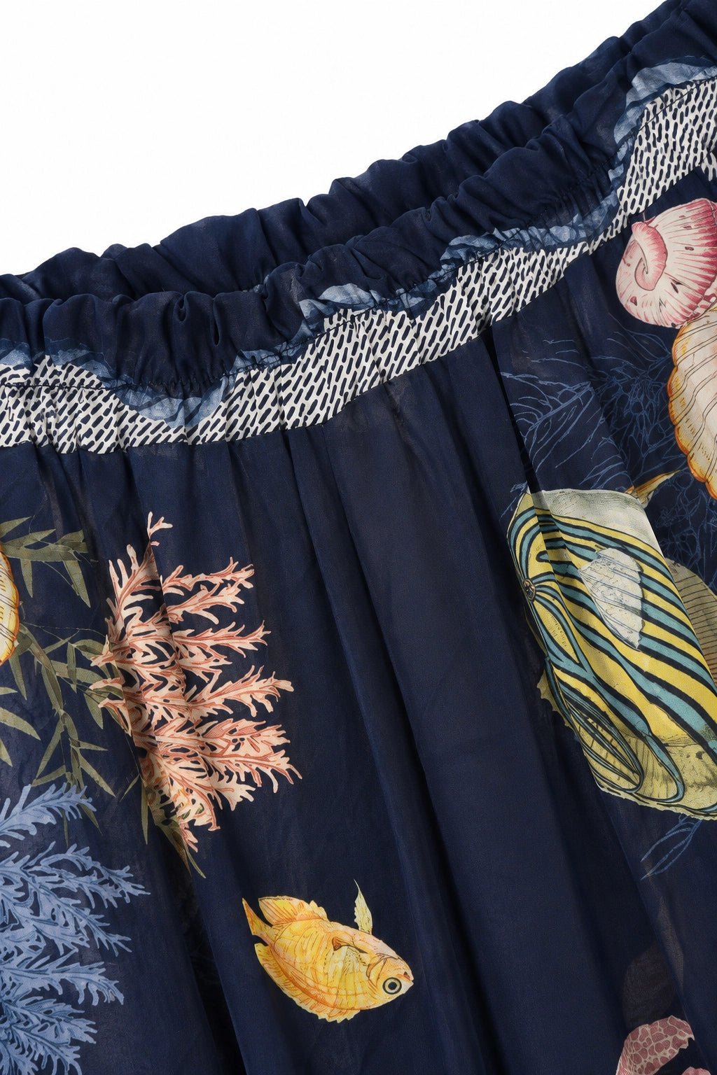 Close-up of a navy blue fabric product, likely a skirt or drape, featuring an ocean-themed print with yellow fish, pink coral, and seashells, finished with a gathered elastic waistband and a white-and-blue patterned trim.
