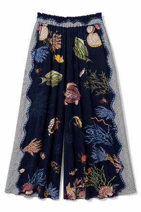A pair of wide-leg cropped pants featuring a dark navy blue base printed with an underwater theme including fish, seashells, and coral, framed by textured gray patterned side panels and a ruffled waistband.