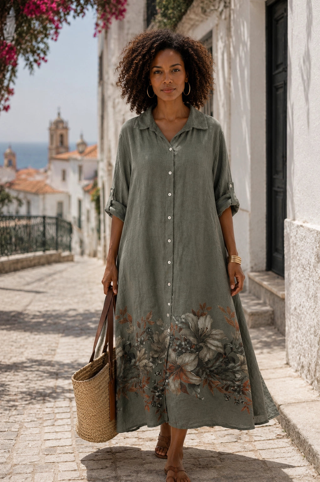 A woman stands on a sunlit cobblestone street wearing a long-sleeve button-front midi dress in muted sage green with a large floral print near the hem. She carries a woven straw tote bag and is posed in front of white buildings with a distant church tower and blue sky.