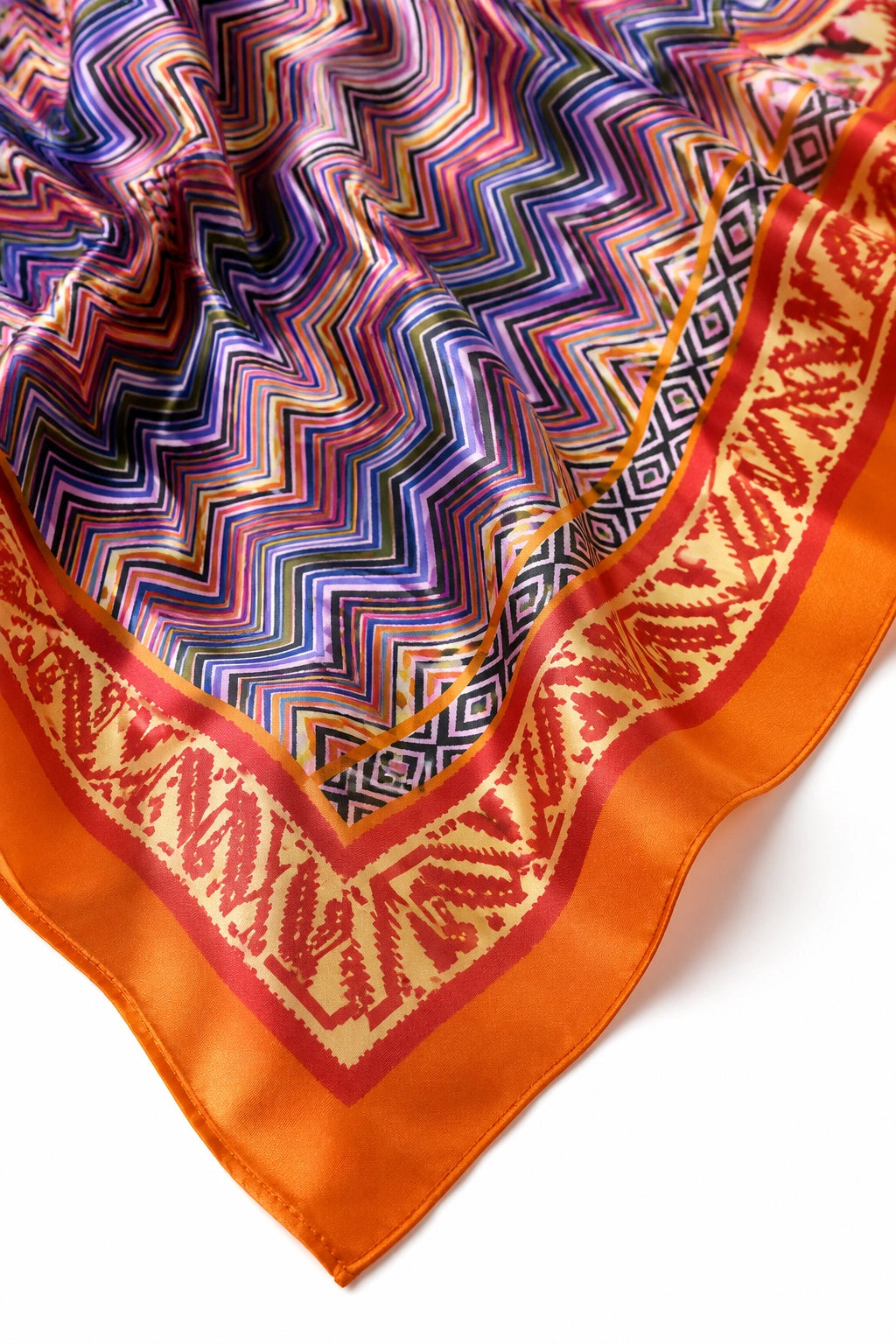 A close-up of a square scarf with a vibrant multicolor zigzag pattern and a wide orange border featuring a red geometric design, laid flat on a white background.