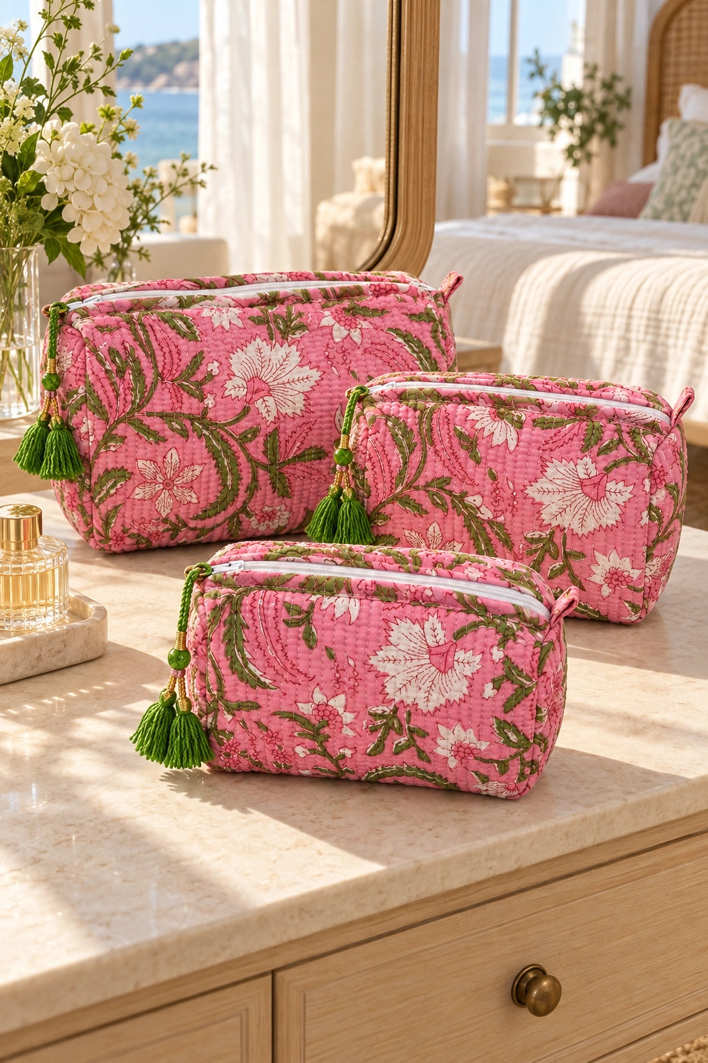 Three quilted cosmetic pouches in a pink floral pattern with green and white flowers and tassel zipper pulls, arranged on a light marble vanity in a sunlit bedroom setting.
