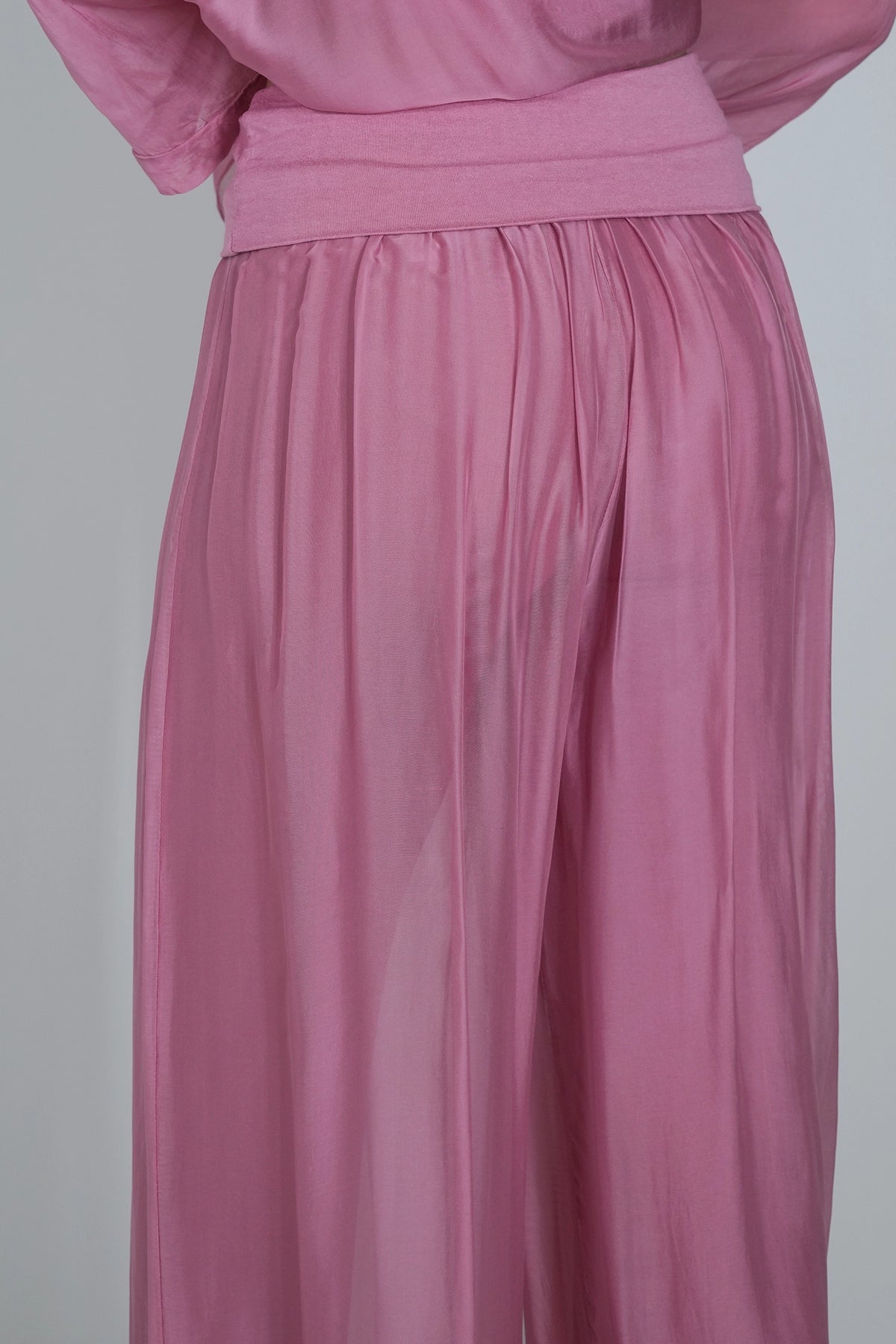 A close-up of a dusty pink garment, showing a wide waistband and flowing pleated fabric, likely a skirt or wide-leg pants, against a plain light gray background.