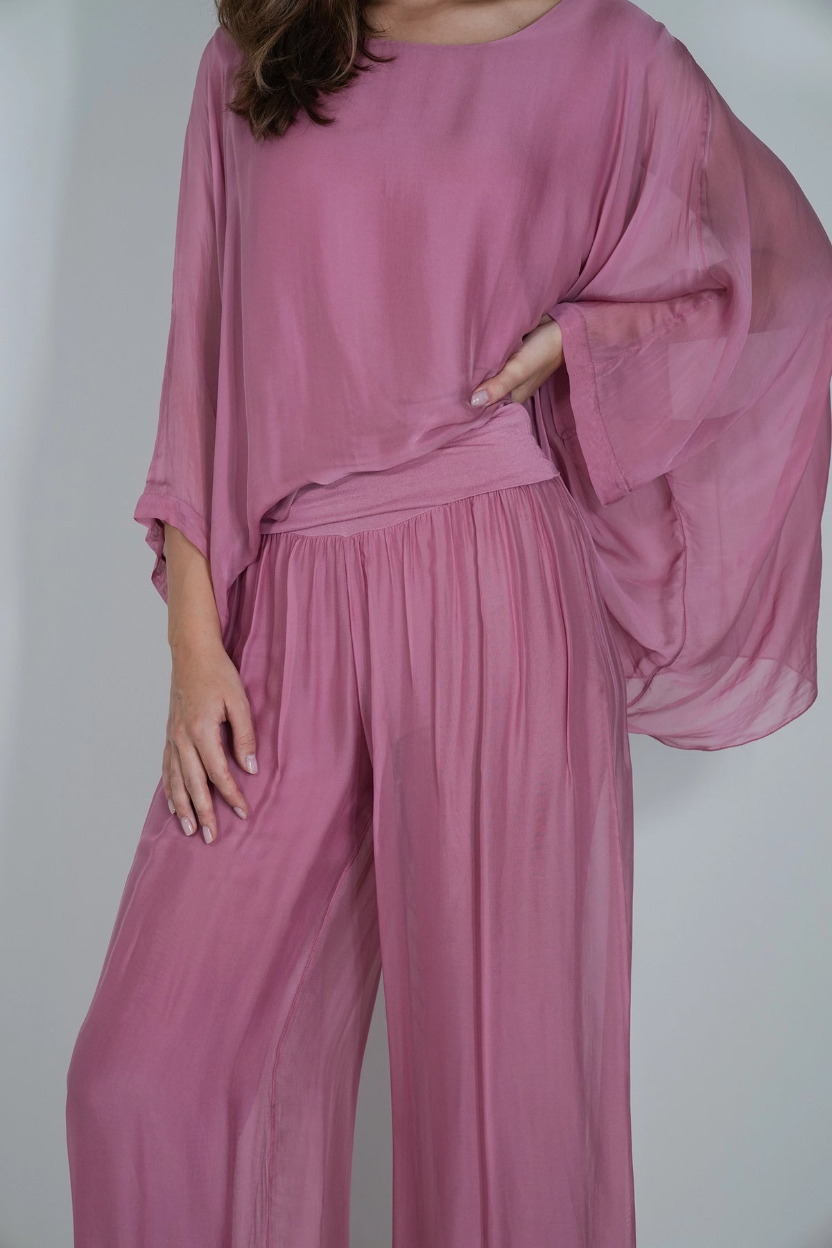 A woman wearing a dusty pink two-piece outfit consisting of a loose, flowy top with wide draped sleeves and matching high-waisted pleated wide-leg pants, shown against a plain light gray background.