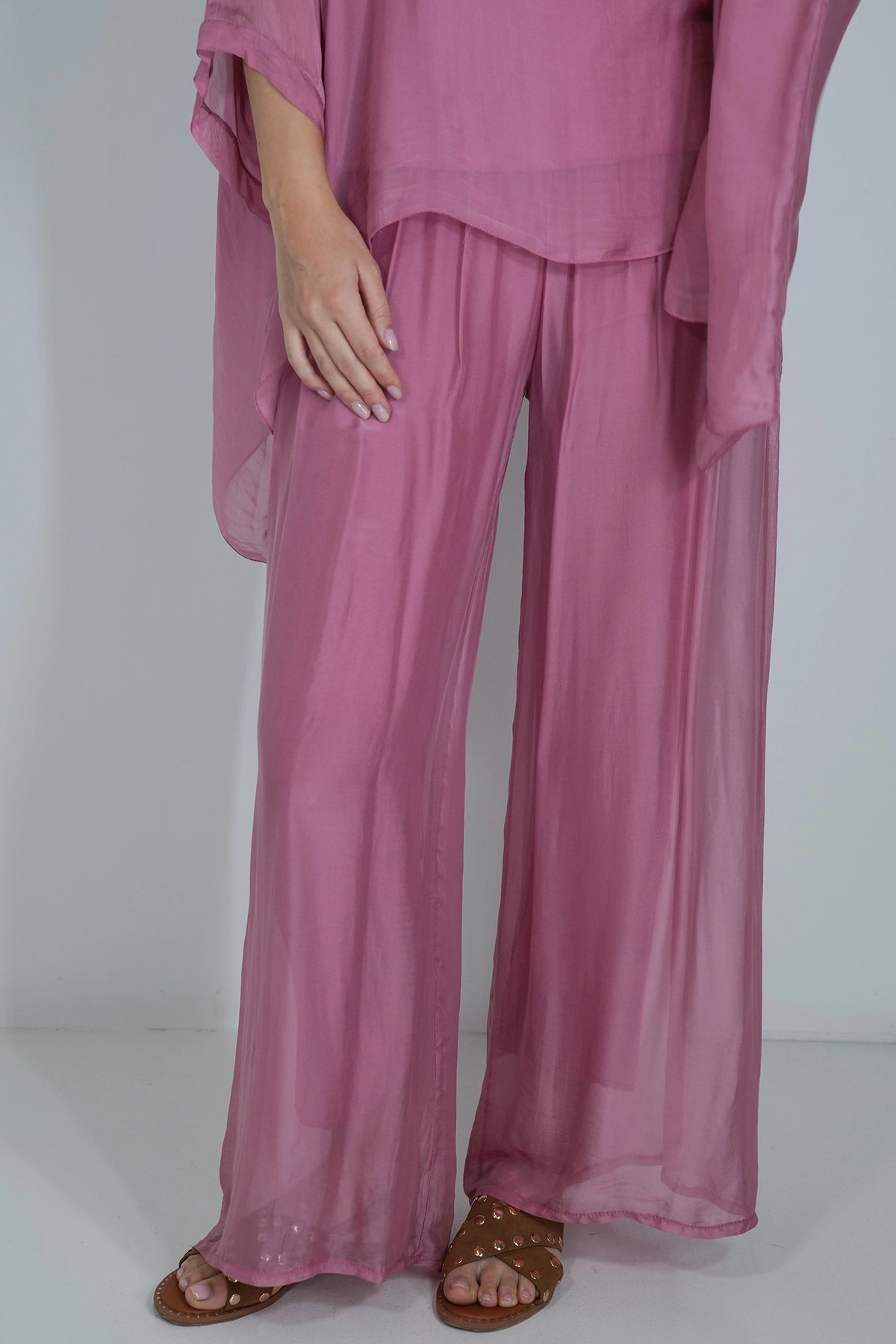A person wearing a dusty pink two-piece outfit with wide-leg sheer pants and a matching loose top, paired with brown studded sandals against a plain light gray studio background.