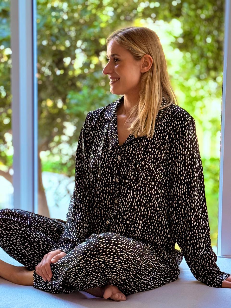 A woman with blonde hair sits cross-legged on a windowsill wearing a black long-sleeve pajama set with a small white floral pattern, looking to the side with a smile. The background shows a bright, sunlit window with green foliage outside.