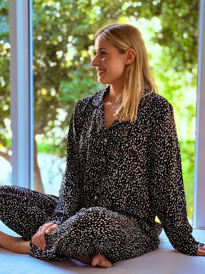 A woman with blonde hair sits cross-legged on a windowsill wearing a black long-sleeve pajama set with a small white floral pattern, looking to the side with a smile. The background shows a bright, sunlit window with green foliage outside.