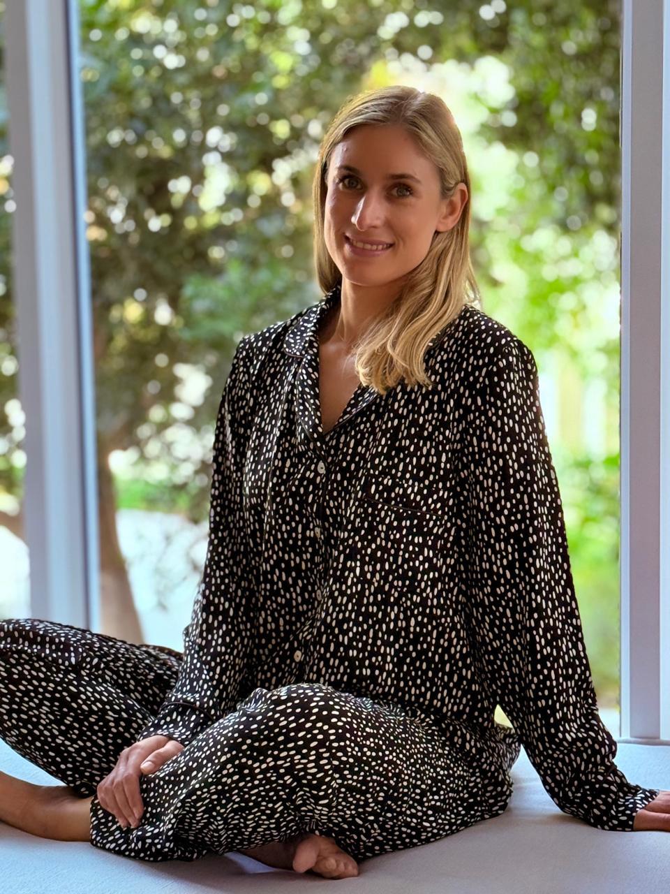 A woman sitting cross-legged in front of a large window, wearing a matching black and white patterned long-sleeve button-up shirt and trousers set.