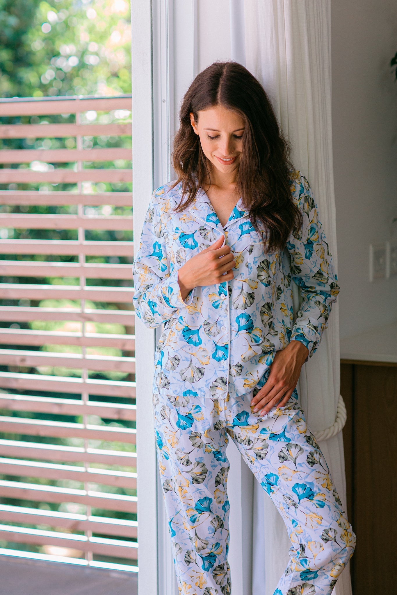 A woman wearing a matching long-sleeve pajama set with a blue and yellow floral print on a white background, standing by a window with white curtains.