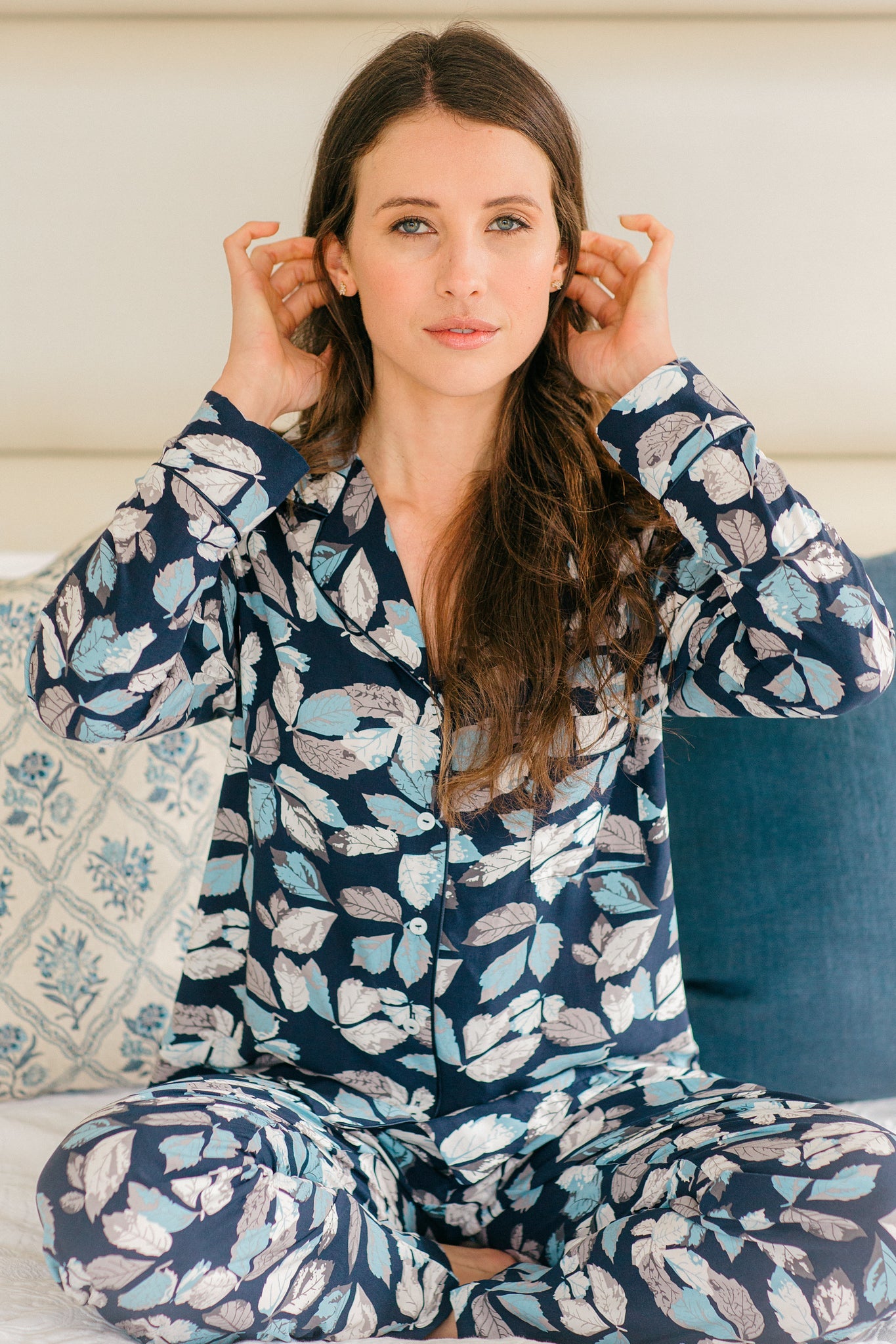 A woman wearing a navy blue pajama set with a light blue and white leaf pattern, sitting on a bed with pillows.