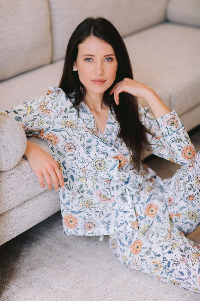 A woman with long dark hair sits on a light beige sofa wearing a matching pajama set with a detailed floral and botanical print in muted blue, orange, and green tones on a white base.