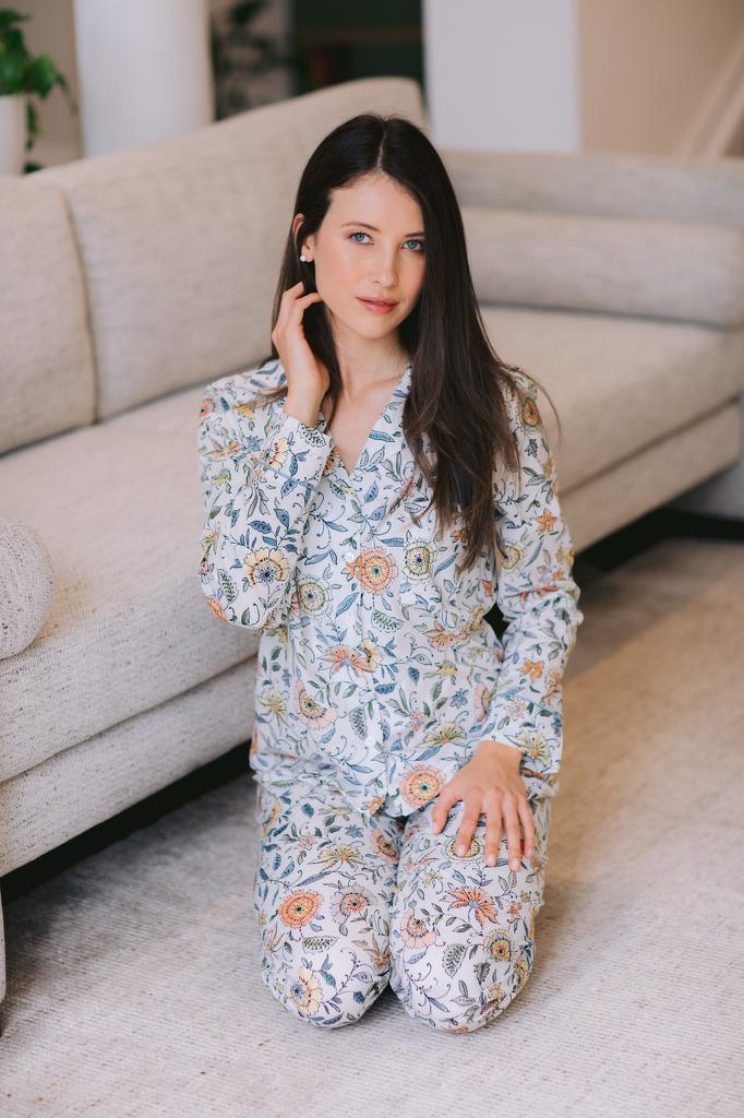 A woman kneeling on a light-colored carpet in front of a beige sofa, wearing a matching long-sleeve pajama set with a blue, orange, and white floral pattern.