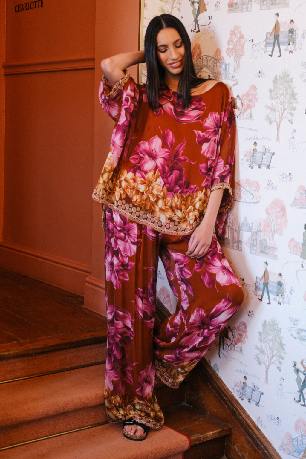 A woman models a rust-brown two-piece pajama set with a vibrant pink and orange floral print, standing on wooden stairs against a patterned wallpaper background.