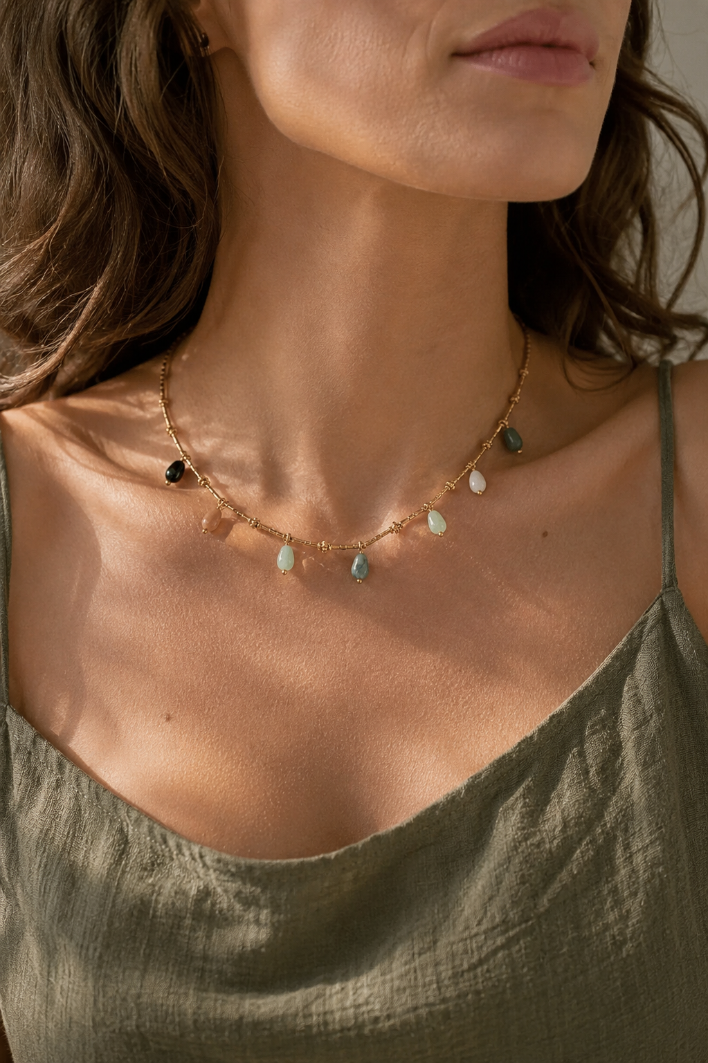 A close-up lifestyle shot of a woman wearing a delicate gold chain necklace adorned with small, irregular natural stone charms in various earthy colors, paired with an olive green top.