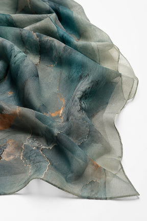 A lightweight sheer scarf with an abstract watercolor pattern in shades of teal, slate blue, and gray, accented with fine gold veining, draped elegantly on a white background.