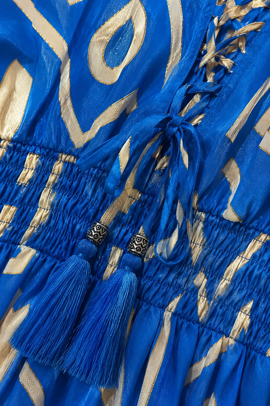 Close-up of a blue and beige patterned garment with a smocked waist, blue drawstring ties, and tassels with silver decorative beads.