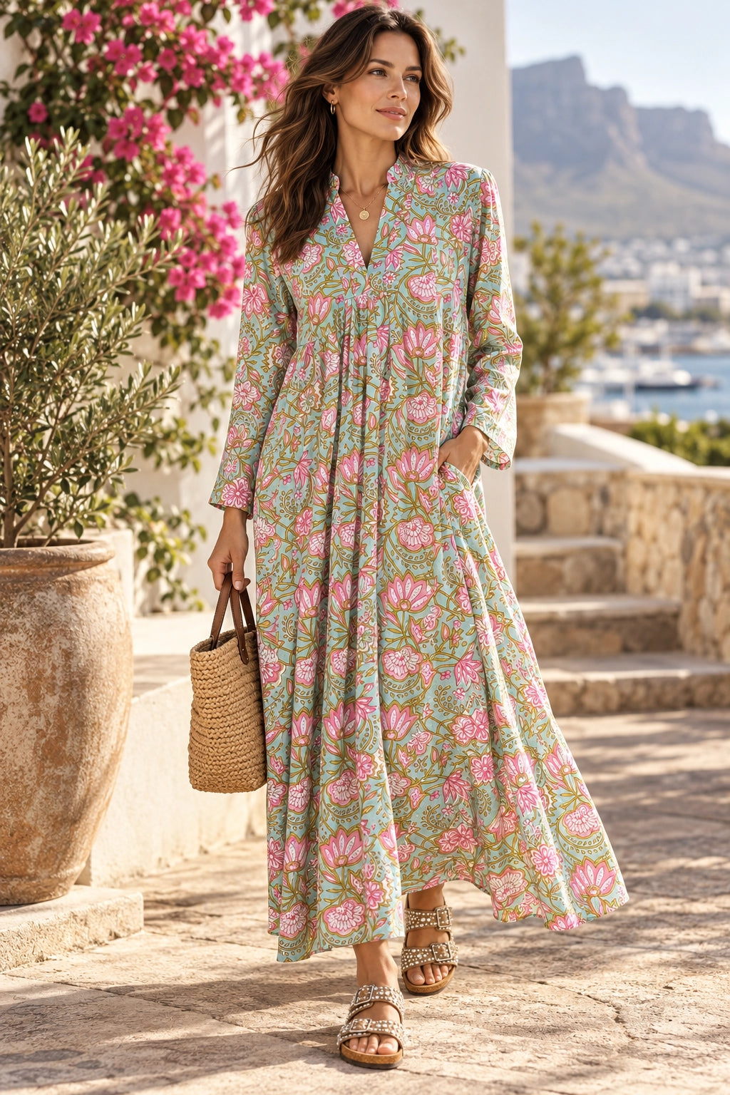 A woman wearing a long-sleeve, V-neck maxi dress with a pink and green floral print, walking outdoors on a stone path next to a large potted plant with pink flowers.