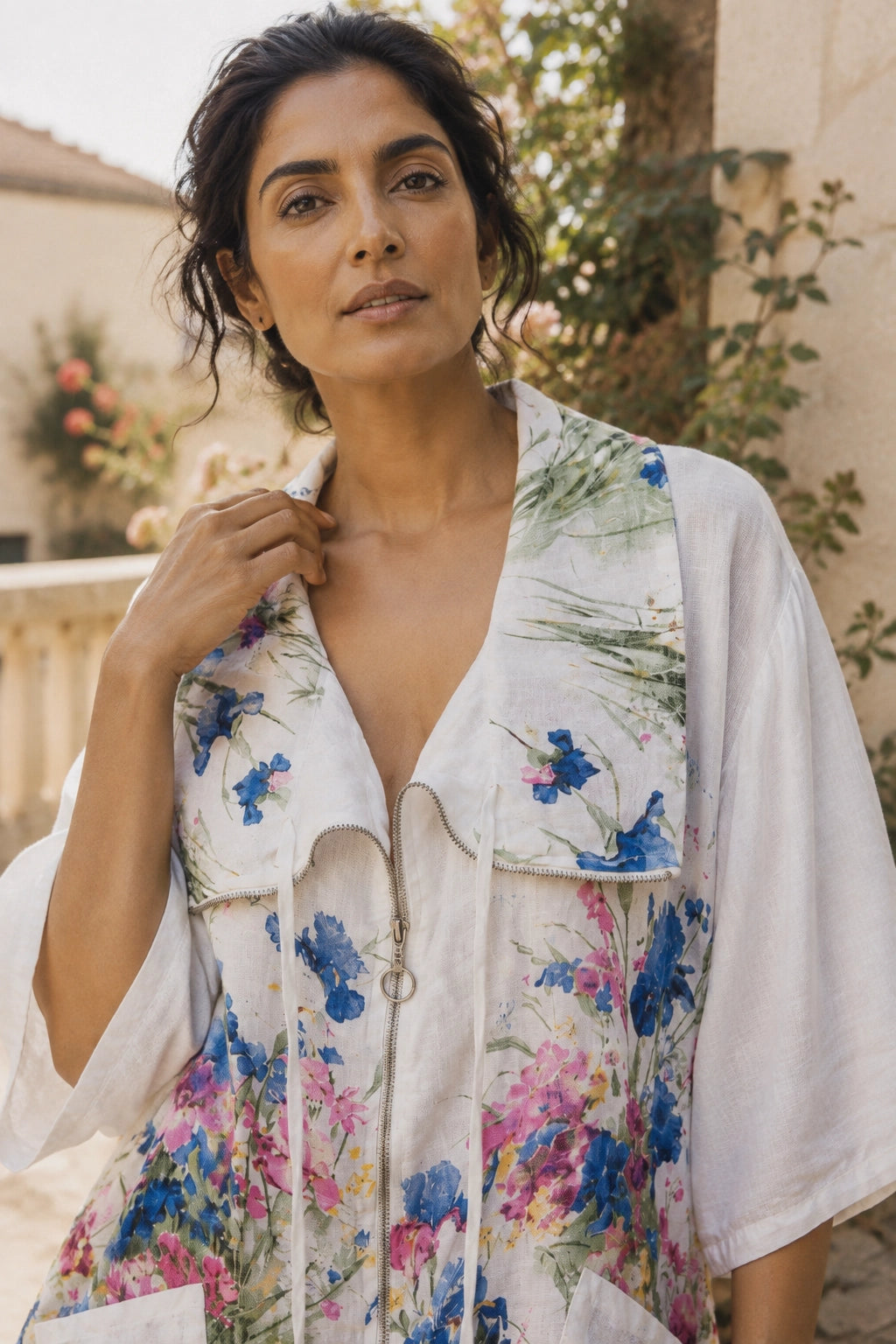 A woman wearing a white kimono-style cover-up with a colorful floral print in blue, pink, and green, photographed outdoors against a warm-toned stone wall and greenery.