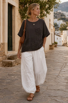 A woman walking down a cobblestone street wearing a dark charcoal gray short-sleeve top with floral embroidery and white wide-leg embroidered pants, carrying a brown shoulder bag.