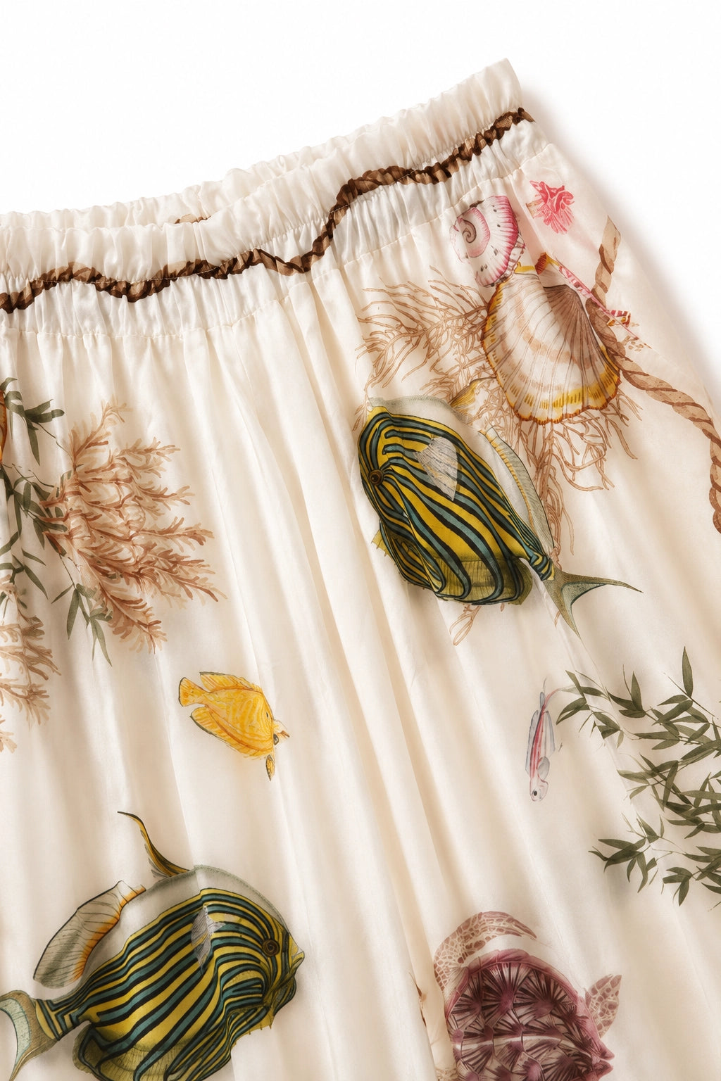 Close-up of a pleated skirt in cream with a colorful underwater print featuring tropical fish, seashells, and seaweed, finished with a brown braided waistband.