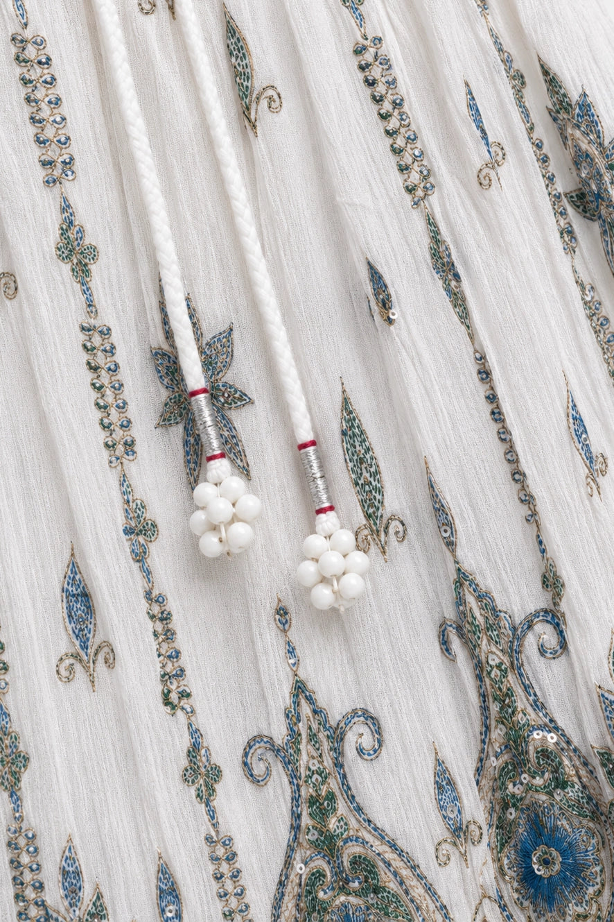 Close-up of a white embroidered fabric with blue and green paisley patterns and white beaded drawstring tassels.