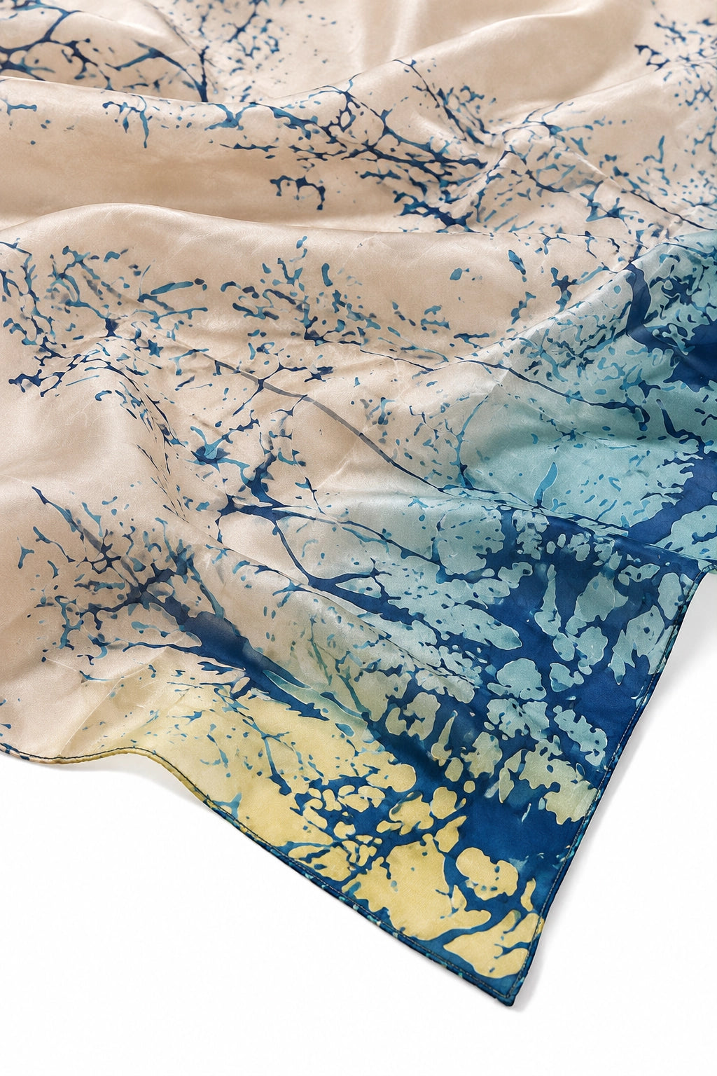 A lightweight silk scarf with a marbled abstract pattern, draped elegantly on a white background. The design features a warm beige base with splashes of deep blue and a soft yellow-gold near the bottom right corner.