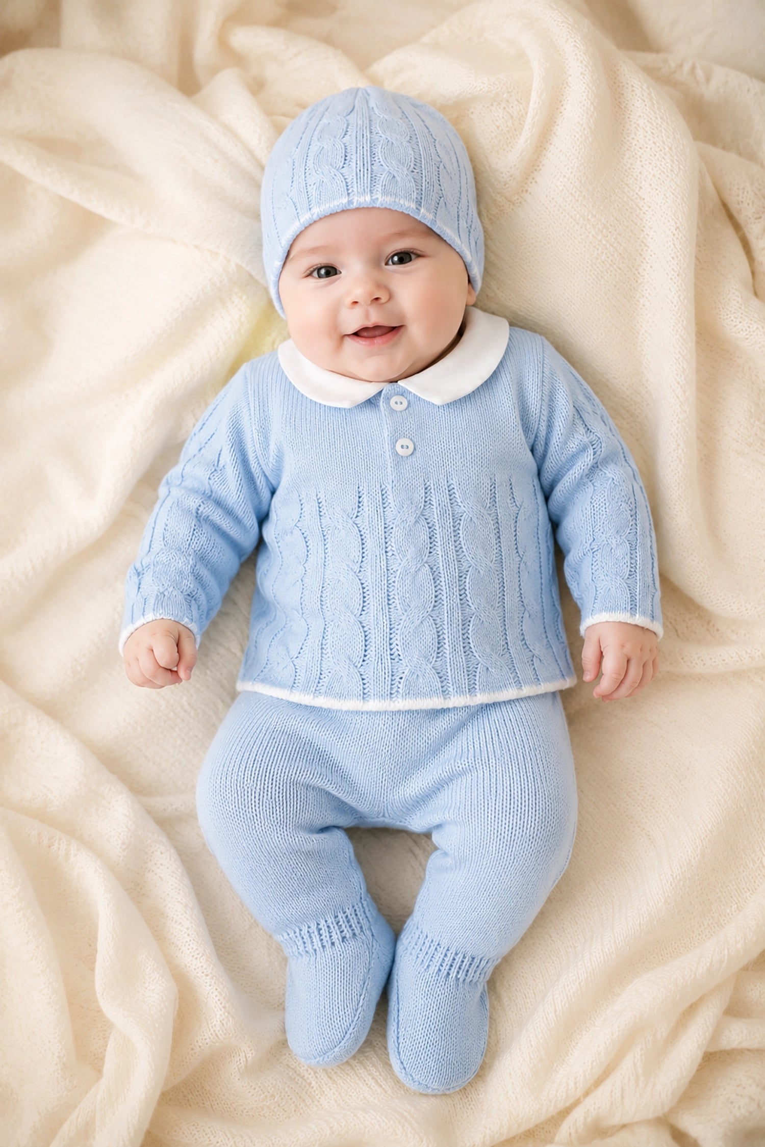 Baby wearing a blue knitted outfit with a matching hat on a soft beige blanket.