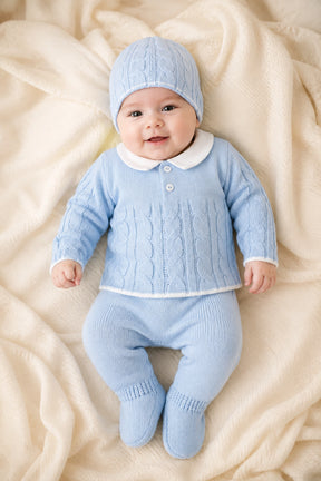 Baby wearing a blue knitted outfit with a matching hat on a soft beige blanket.