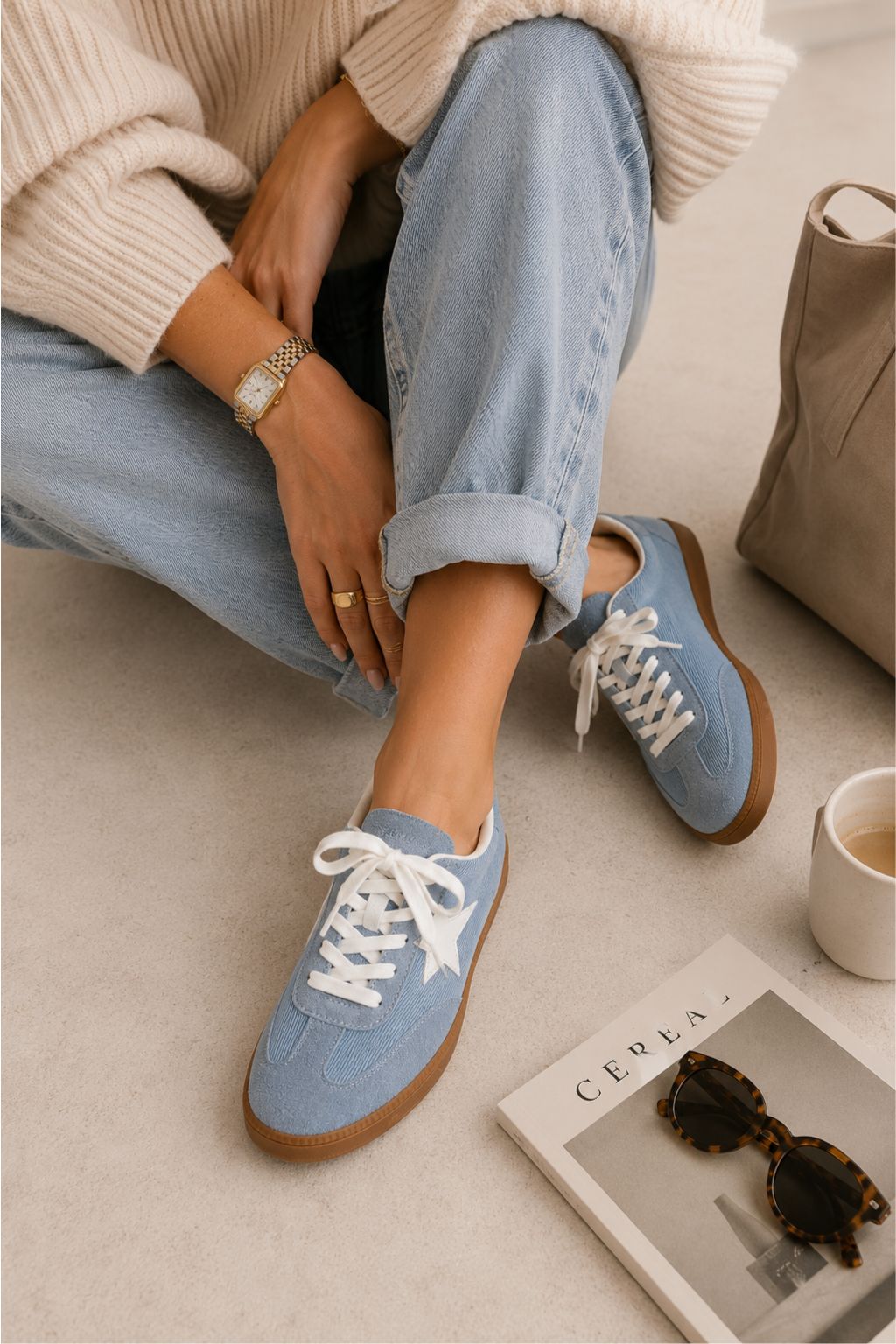 Person wearing blue sneakers with white laces, sitting on a couch with a cup and sunglasses.
