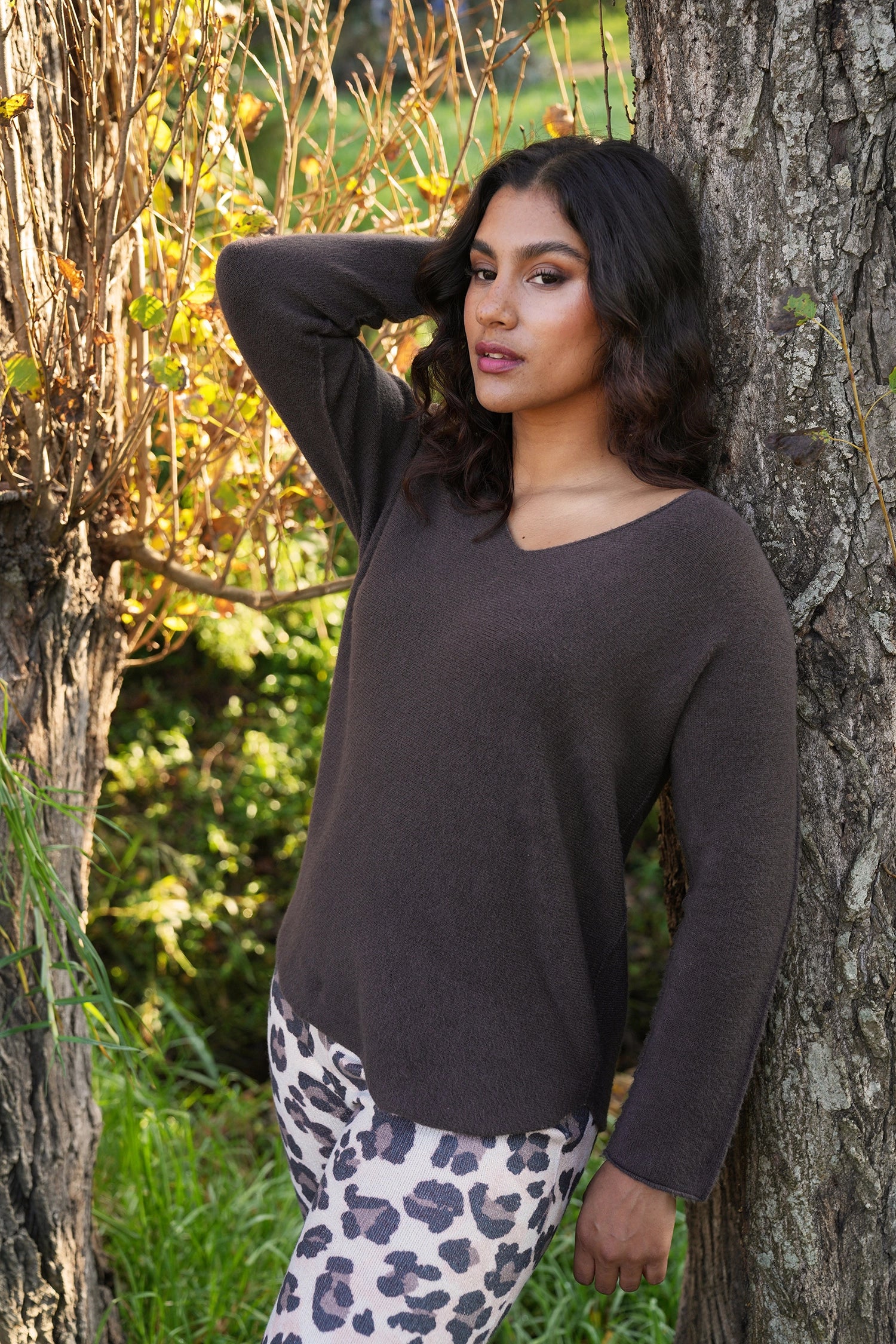 Woman in a brown sweater and patterned pants leaning against a tree in an outdoor setting