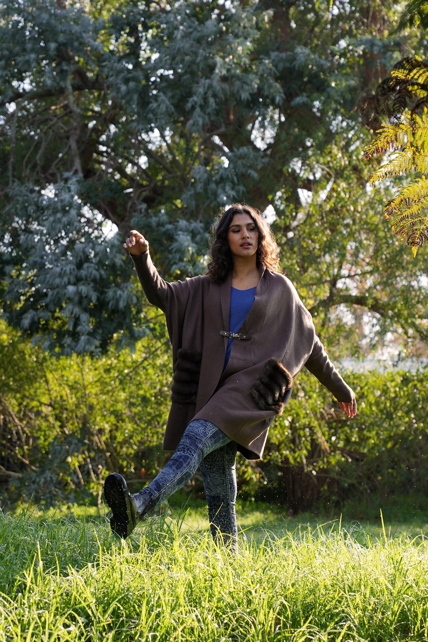 Woman in a brown coat and blue jeans running through a grassy field with trees in the background