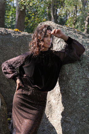 Woman in a dark outfit standing against a stone wall with trees in the background