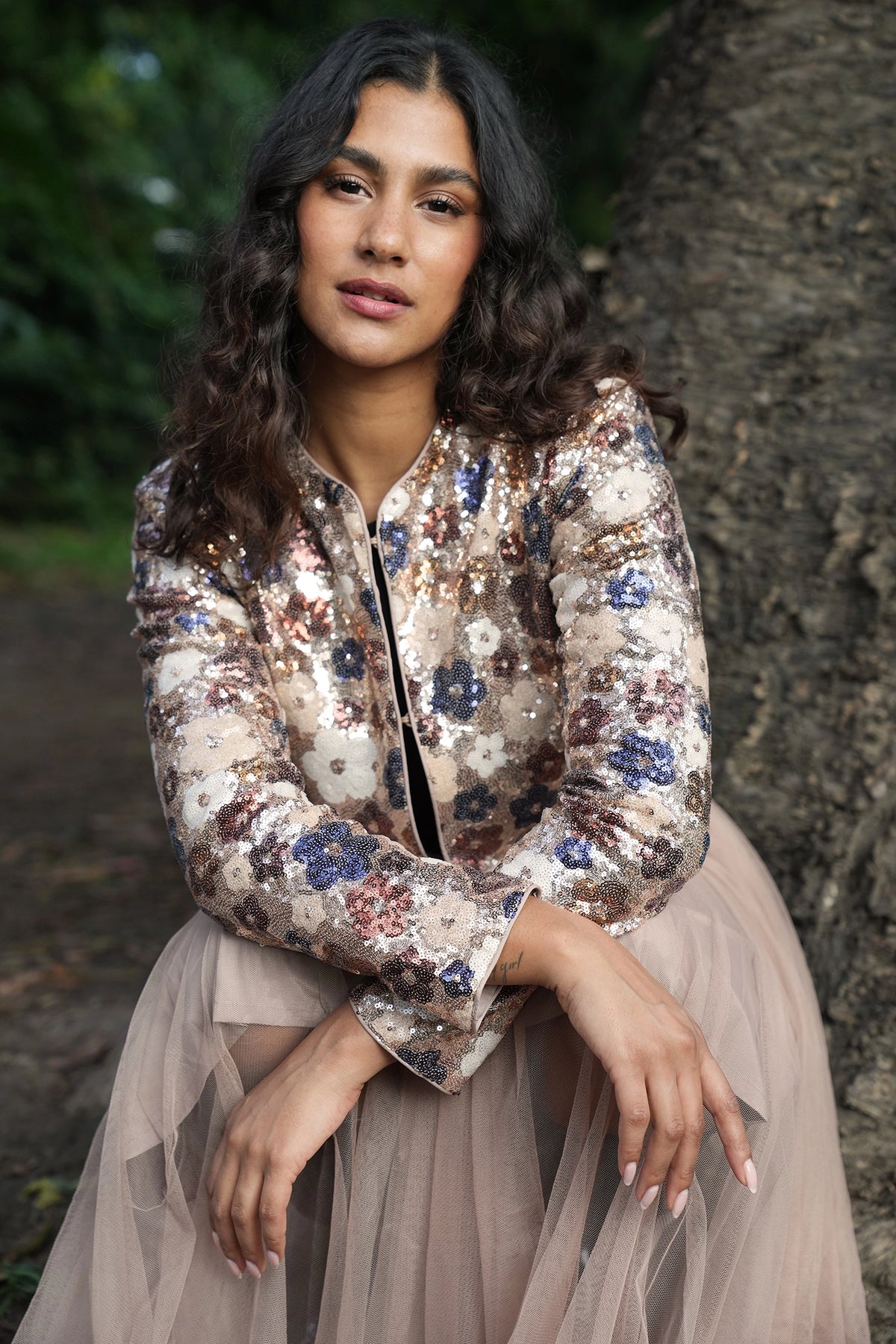 Woman wearing a sequined top and tulle skirt sitting against a tree.