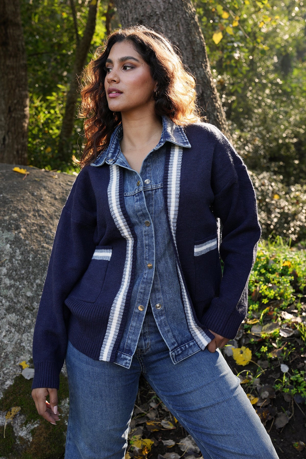 Woman wearing a blue cardigan with white trim standing outdoors near trees and rocks.
