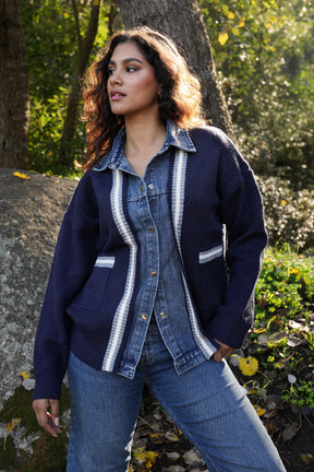 Woman wearing a blue cardigan with white trim standing outdoors near trees and rocks.