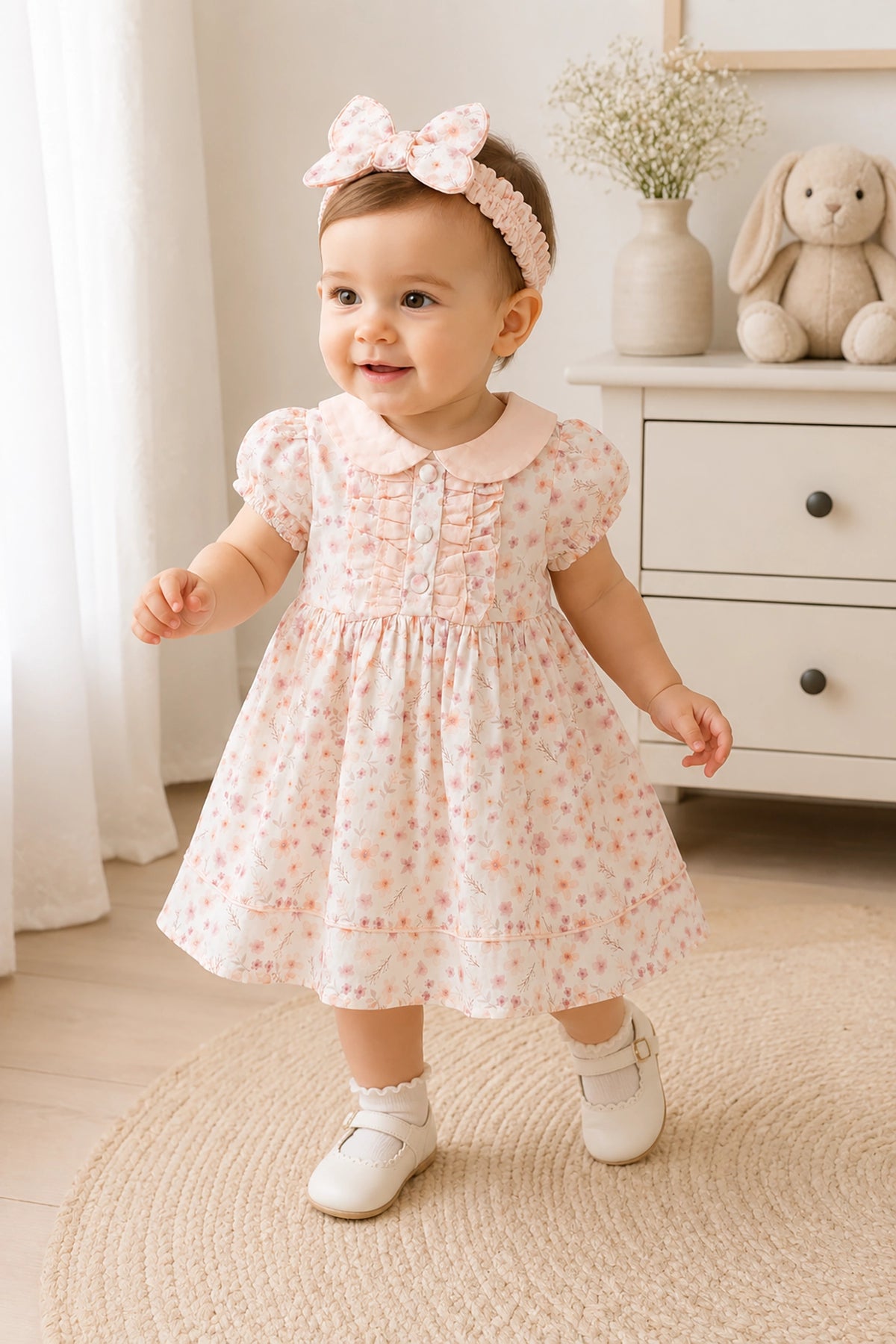 Baby girl in a floral dress standing in a room with a white dresser and decorative items.