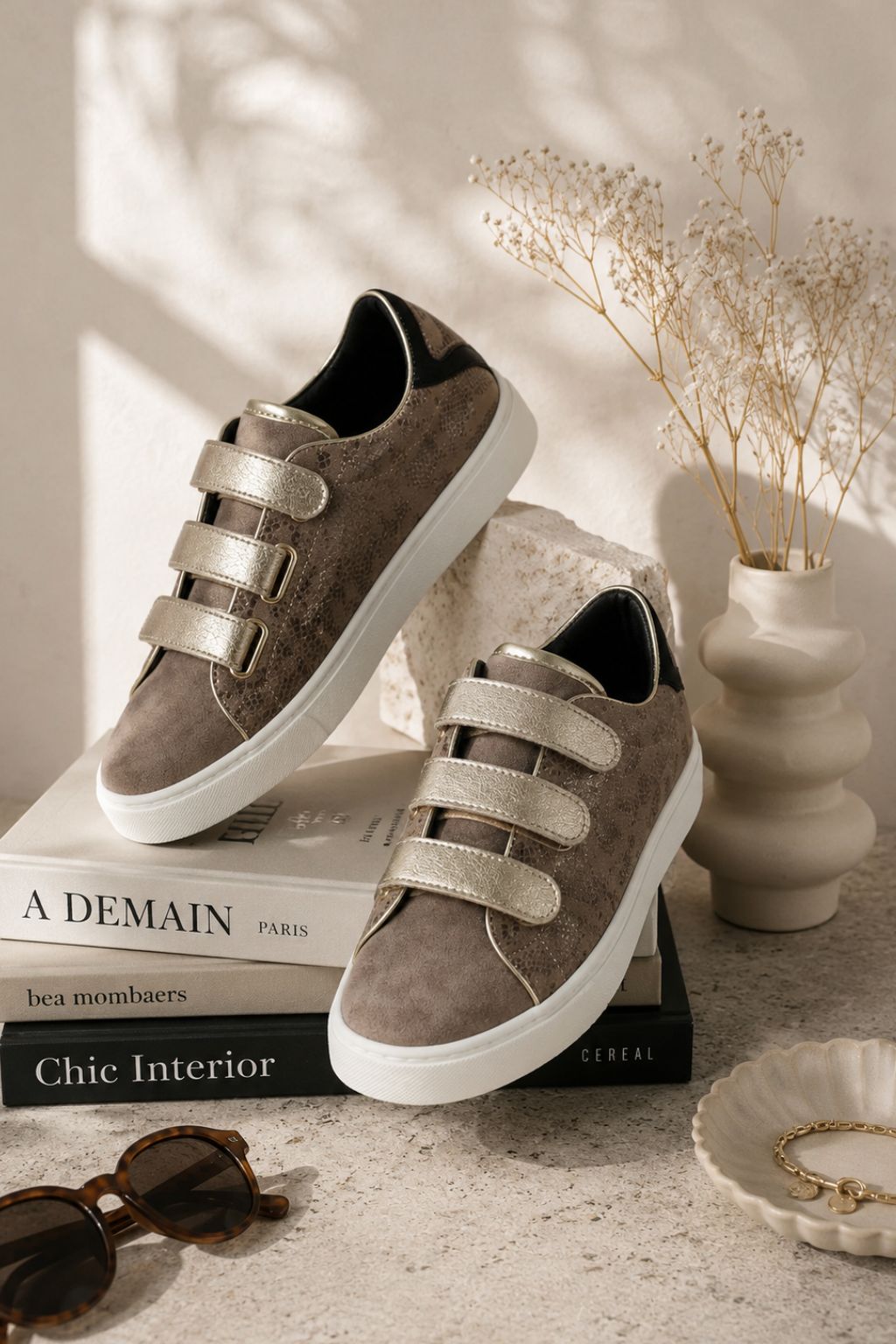 Pair of brown sneakers with velcro straps on a stack of books, with a vase and decor items in the background.