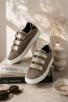 Pair of brown sneakers with velcro straps on a stack of books, with a vase and decor items in the background.