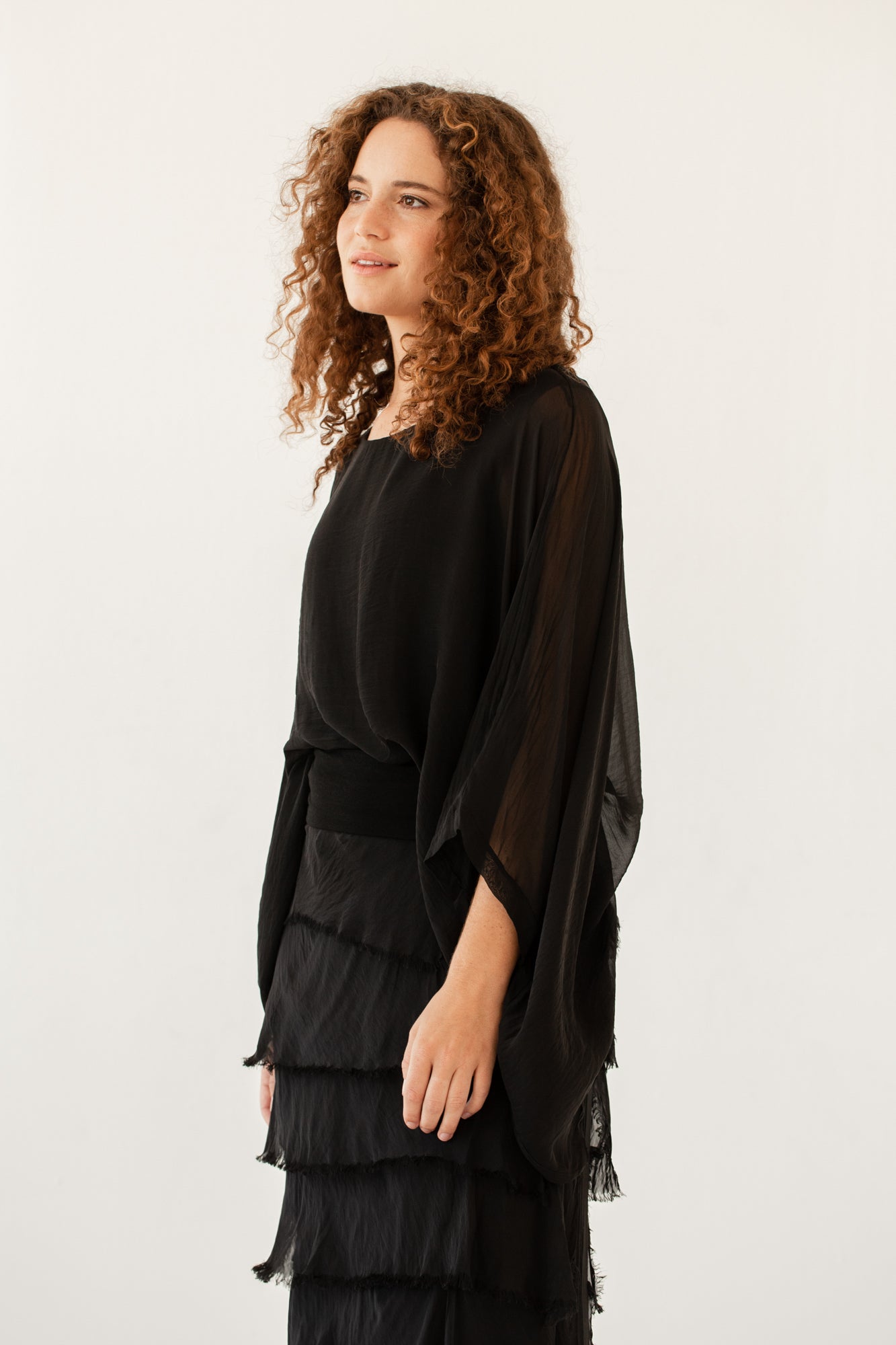Model wearing black silk poncho top with sheer draped sleeves, side view